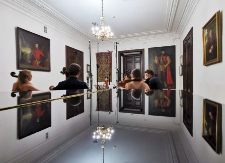 A photograph. A string concert in Villa Decius. The musicians play the violin and the cello in the Ball Room at Villa Decius. The silhuettes reflect in a gloss black surface of the piano in the foreground, in the bottom of the frame.